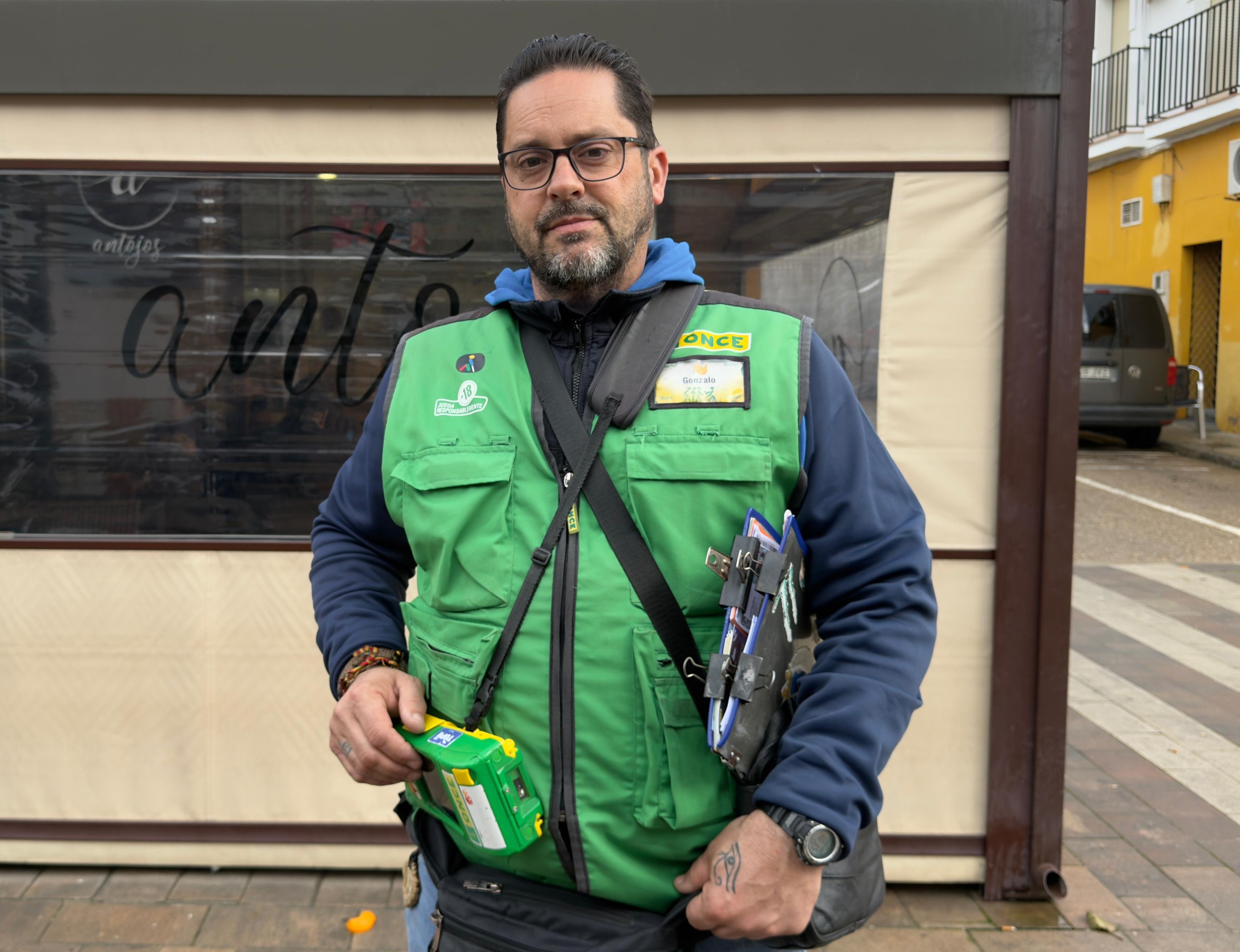 Gonzalo Sánchez Aguilar lleva el chaleco verde, que le identifica como agente vendedor de los productos de juego de la ONCE, y porta el TPV y una carpeta con cupones y resto de productos. Imagen plano medio