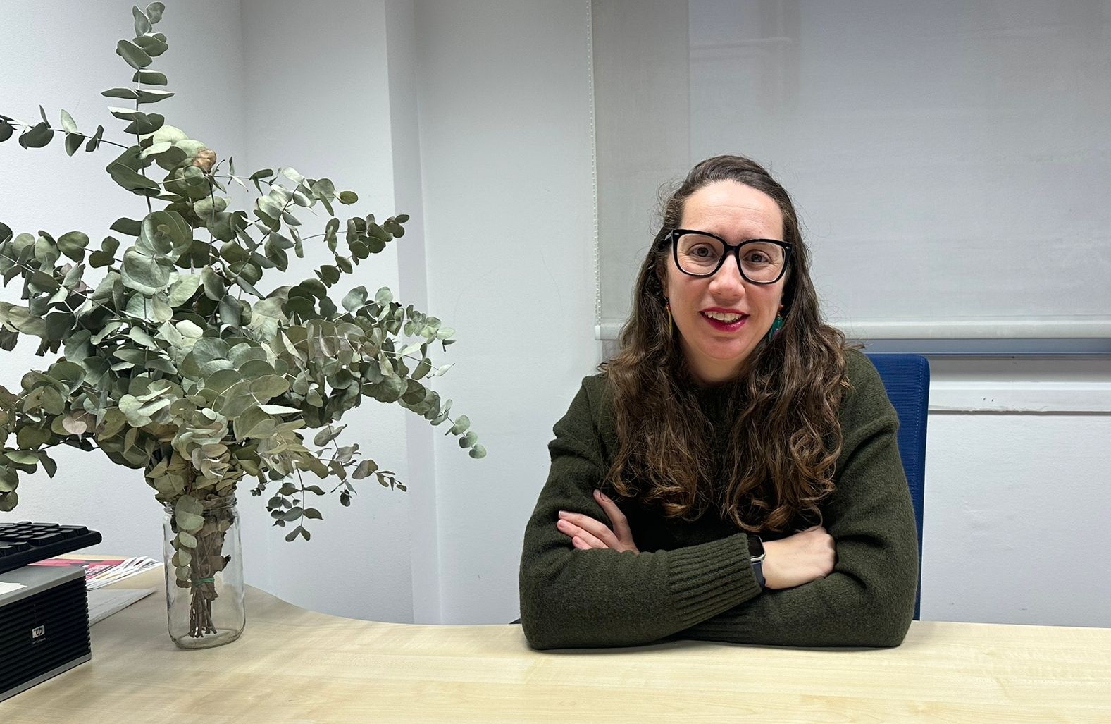 Andrea Ríos Rodríguez posa sonriente en su puesto de trabajo en Inserta Empleo de Fundación ONCE, en la mesa tiene un florero con hojas de eucalipto