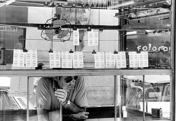 Un kiosco primitivo de la ONCE, con el vendedor dentro y varios cupones colgados en el cristal. Fotografía en blanco y negro.