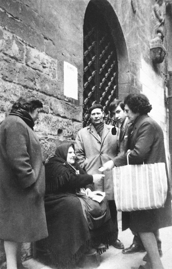 Una mujer pidiendo la caridad en la puerta de una iglesia mientras una familia la atiende y ayuda. Fotografía en blanco y negro