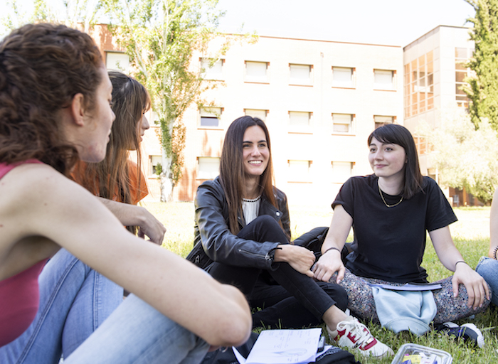 Un grupo de chicas jóvenes sentadas en el césped, en un día soleado, con una universidad al fondo. Llevan comida en recipientes de plástico y cuadernos. Sonríen.