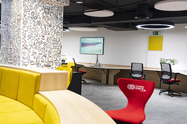 Vista del interior de la sala Espacia, en Fundación ONCE, con un sofá amarillo y circular en primer plano, y mesas y sillas de oficina y de formas curvas al fondo