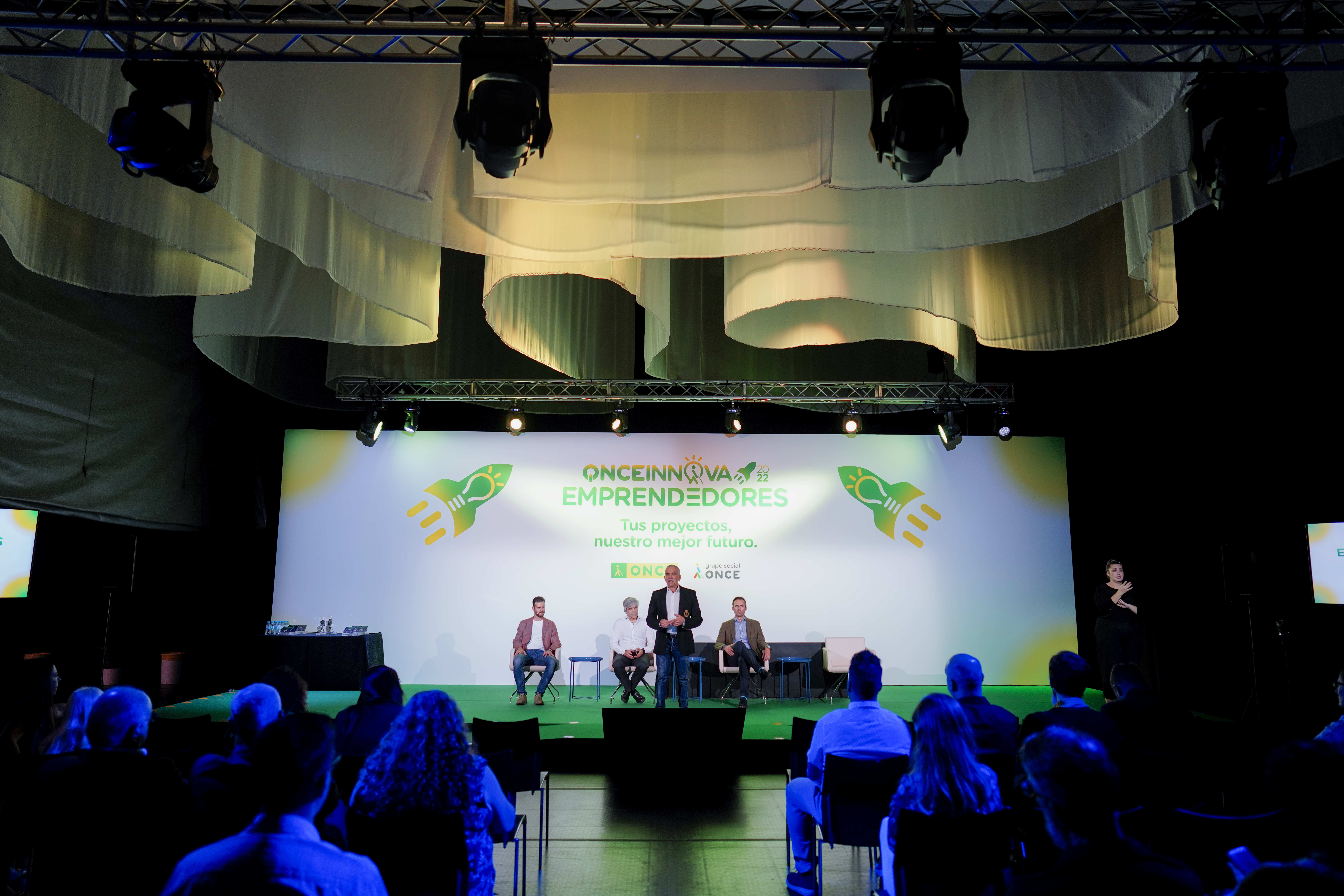Foto frontal del escenario de ONCE Innova Emprendedores 2022, imagen desde el público con focos apuntando al escenario y una nube de telas en el techo.