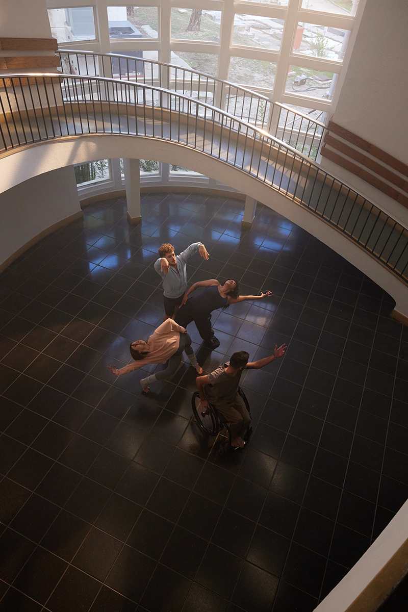 Cuatro personas bailando en el centro de un hall con unas escaleras caracol a su alrededor.  Los bailarines están en línea y uno de ellos va en silla de ruedas