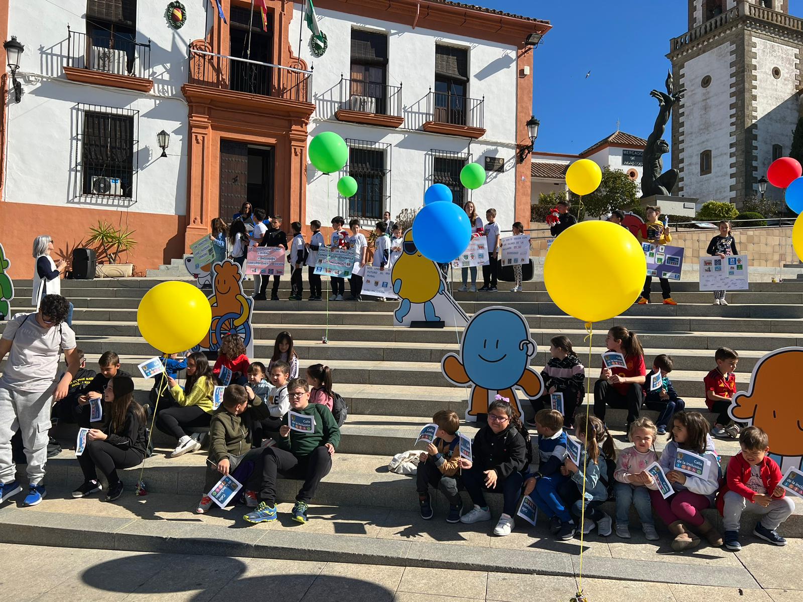 Niños y familias en una escalera, celebrndo con globos y muñecos troquelados en cartón la inauguración de la Calle de la Inclusión