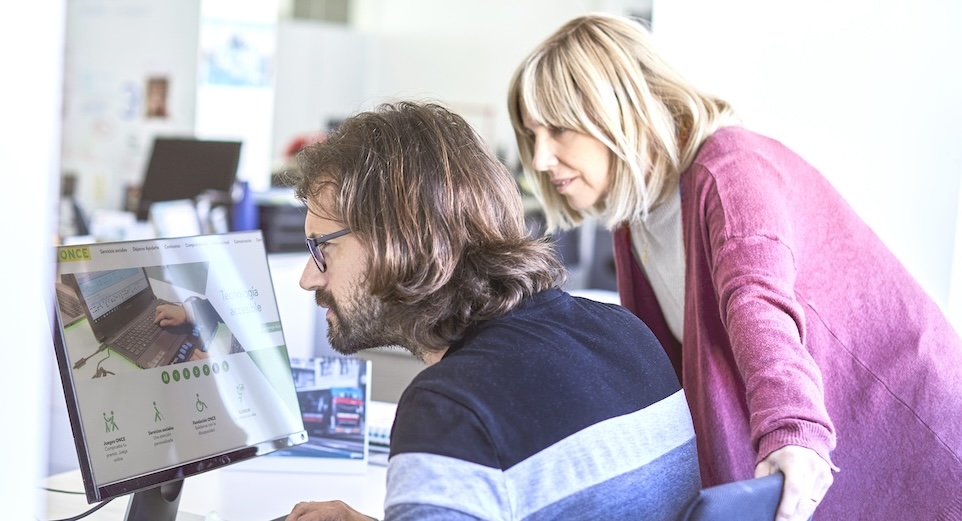 Dos personas delante de un ordenador portátil en la oficina, una con discapacidad visual