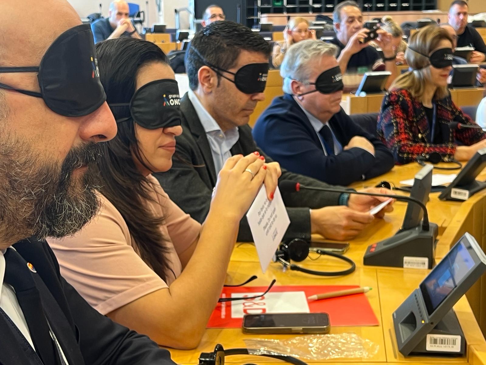 Eurodiputados en sus escaños con antifaz y tocando papeletas de colores