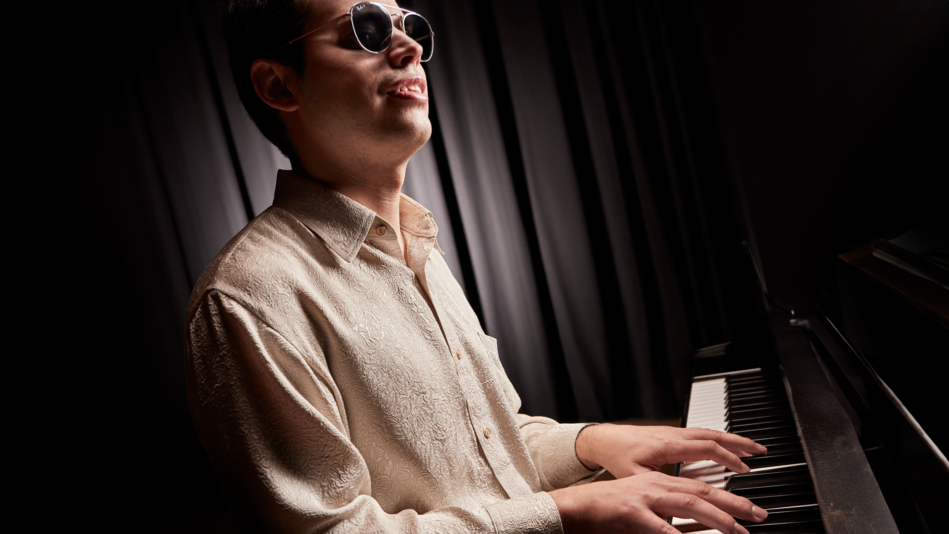 Una persona ciega, con gafas de sol negras y redondeadas, tocando un piano en un entorno oscuro y teatral