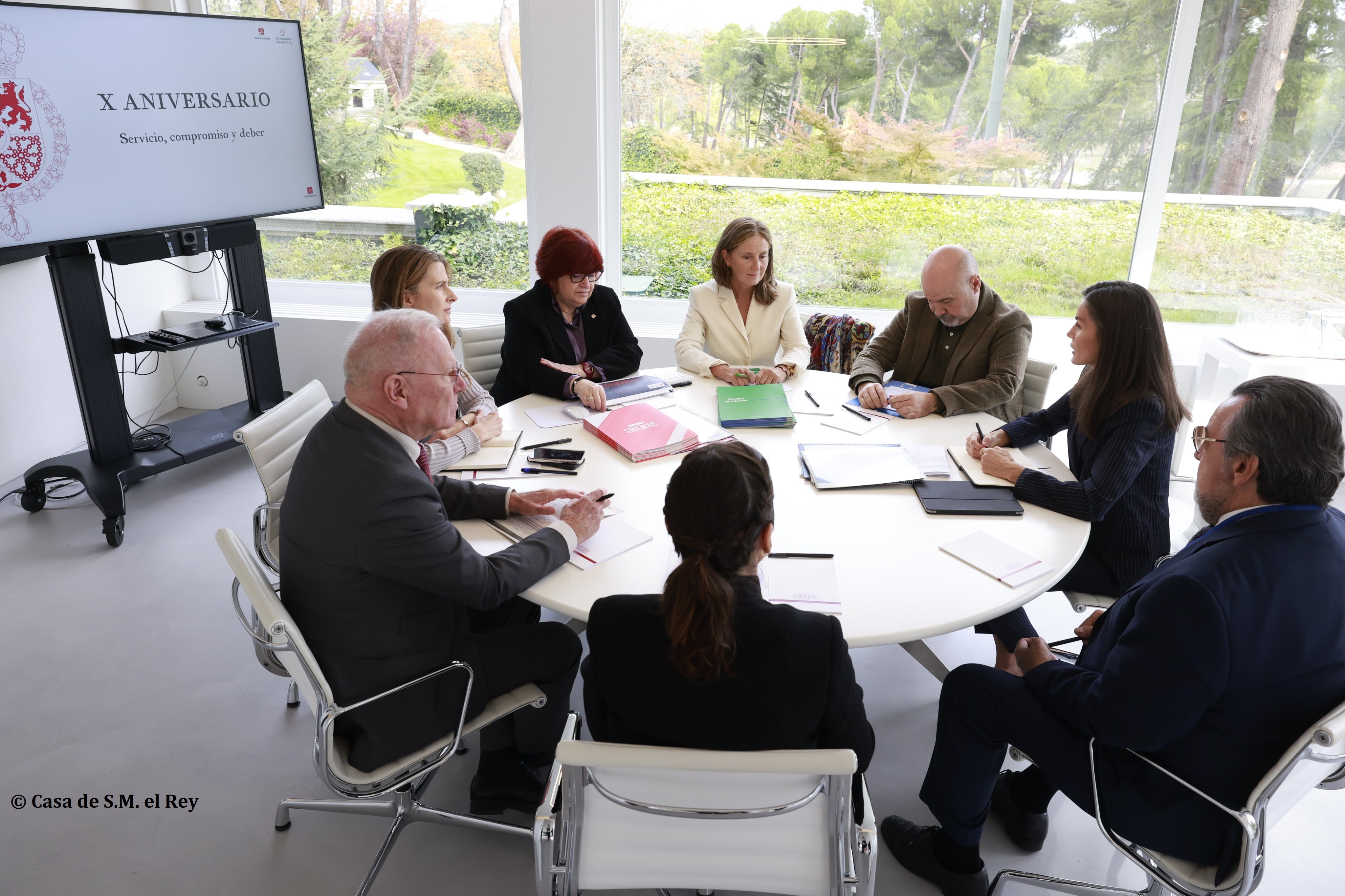 La mesa de trabajo, con la Reina Letizia y Miguel Carballeda además de otros representantes del Tercer Sector, ante una pantalla con diapositiva "X aniversario: Servicio, compromiso y deber"