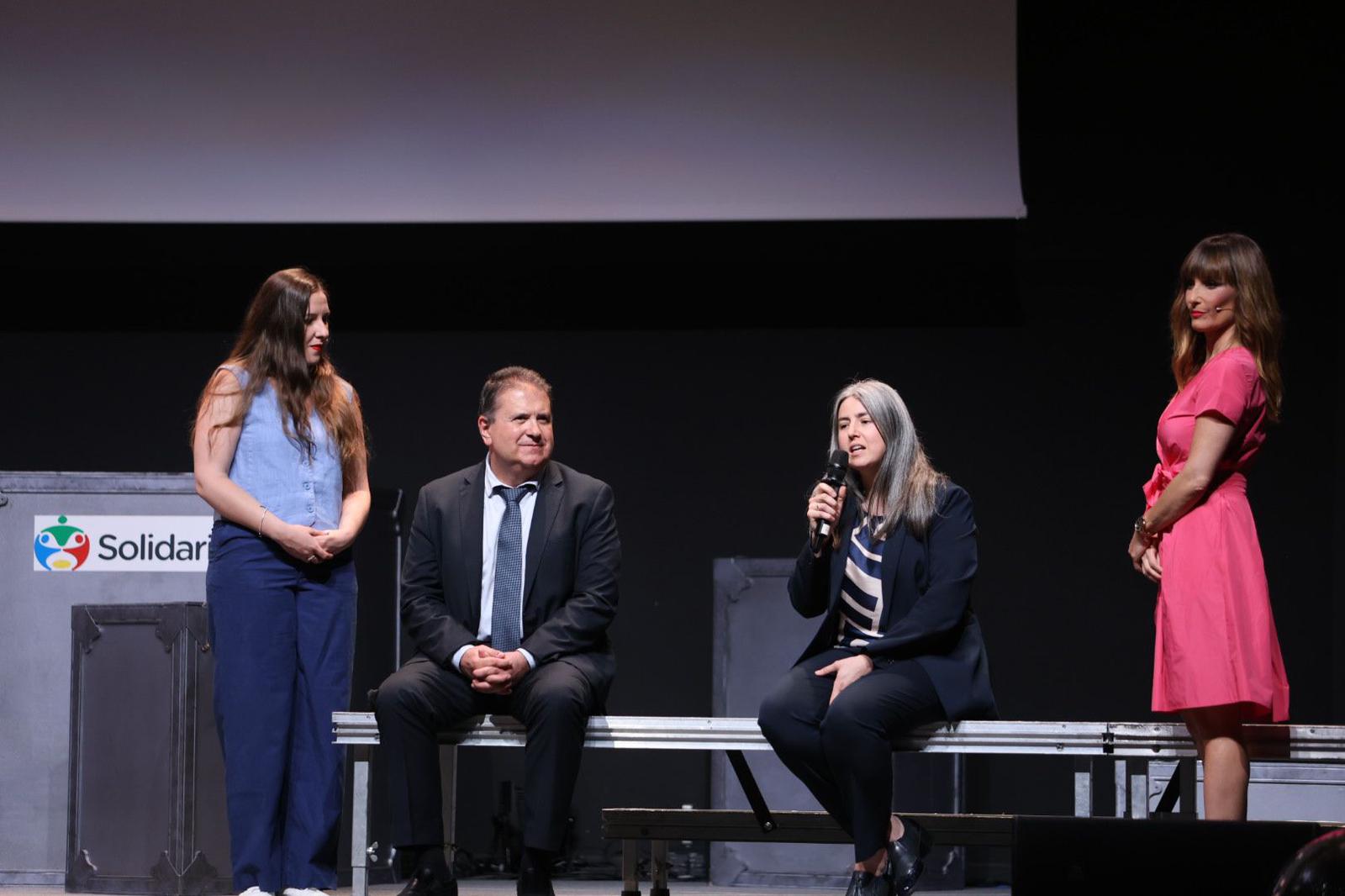 Las presentadoras, de pie, hablan con dos de las autoridades, sentados