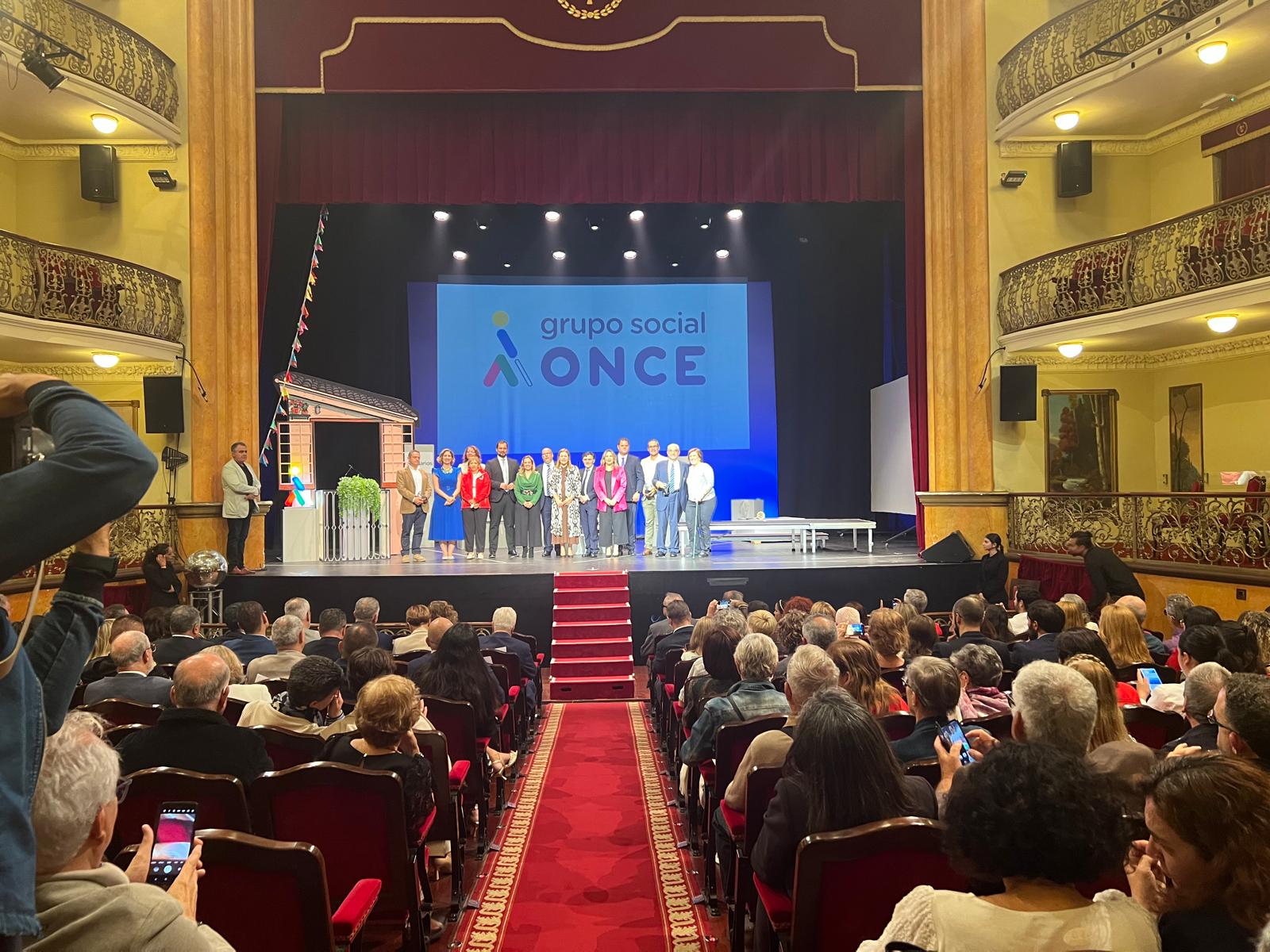 Foto desde la última fila del teatro con todos los premiados y autoridades en el escenario, en la pantalla del fondo un gran logo del Grupo Social ONCE