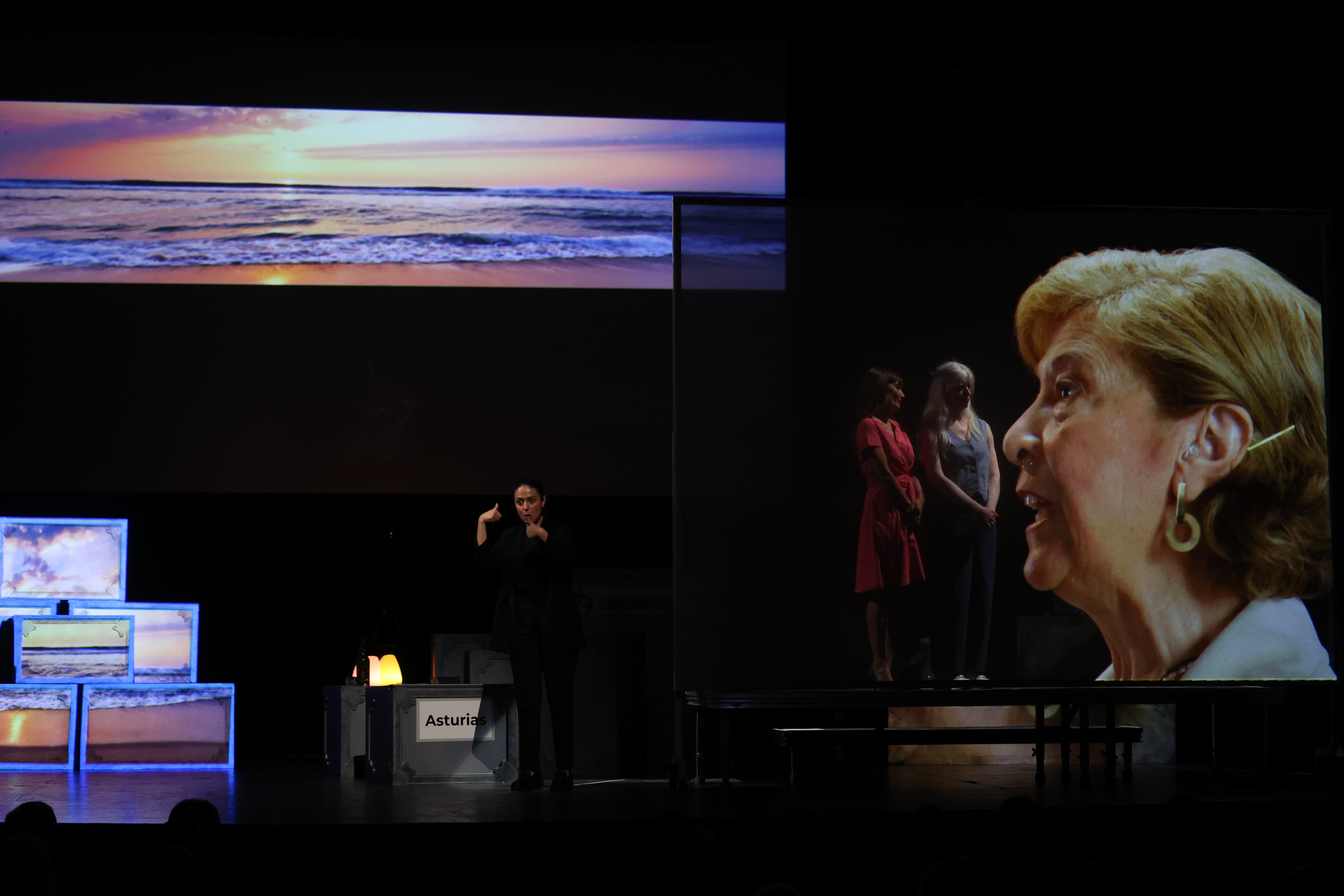 Patty y Laura sobre una plataforma en el escenario del Teatro Filarmónica de Oviedo mirando hacia una pantalla transparente en la que se está proyectando a una mujer hablando sobre la sordoceguera