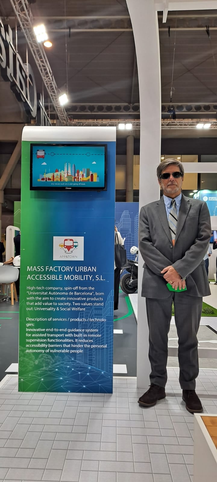 La imagen muestra a Jordi, vestido de manera formal con gafas de sol y de pie,  al lado del stand promocional de App&Town Companion. El panel central destaca con colores degradados en azul y verde, junto con el logotipo de la aplicación, que incluye un ícono de transporte público. En la parte superior del panel hay una pantalla mostrando una animación de una ciudad moderna con edificios y un vehículo de transporte, reforzando el tema de movilidad urbana. En el texto del panel, se describe a Mass Factory Urban Accessible Mobility, S.L., la empresa responsable de la solución, como una spin-off de la Universitat Autònoma de Barcelona, comprometida con la innovación tecnológica, el bienestar social y la accesibilidad.