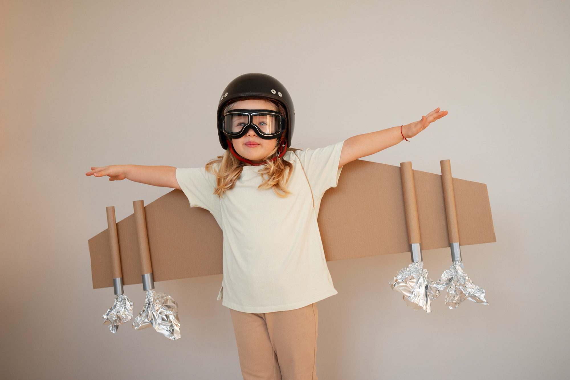Una niña jugando con alas de cartón y casco de piloto de aviones