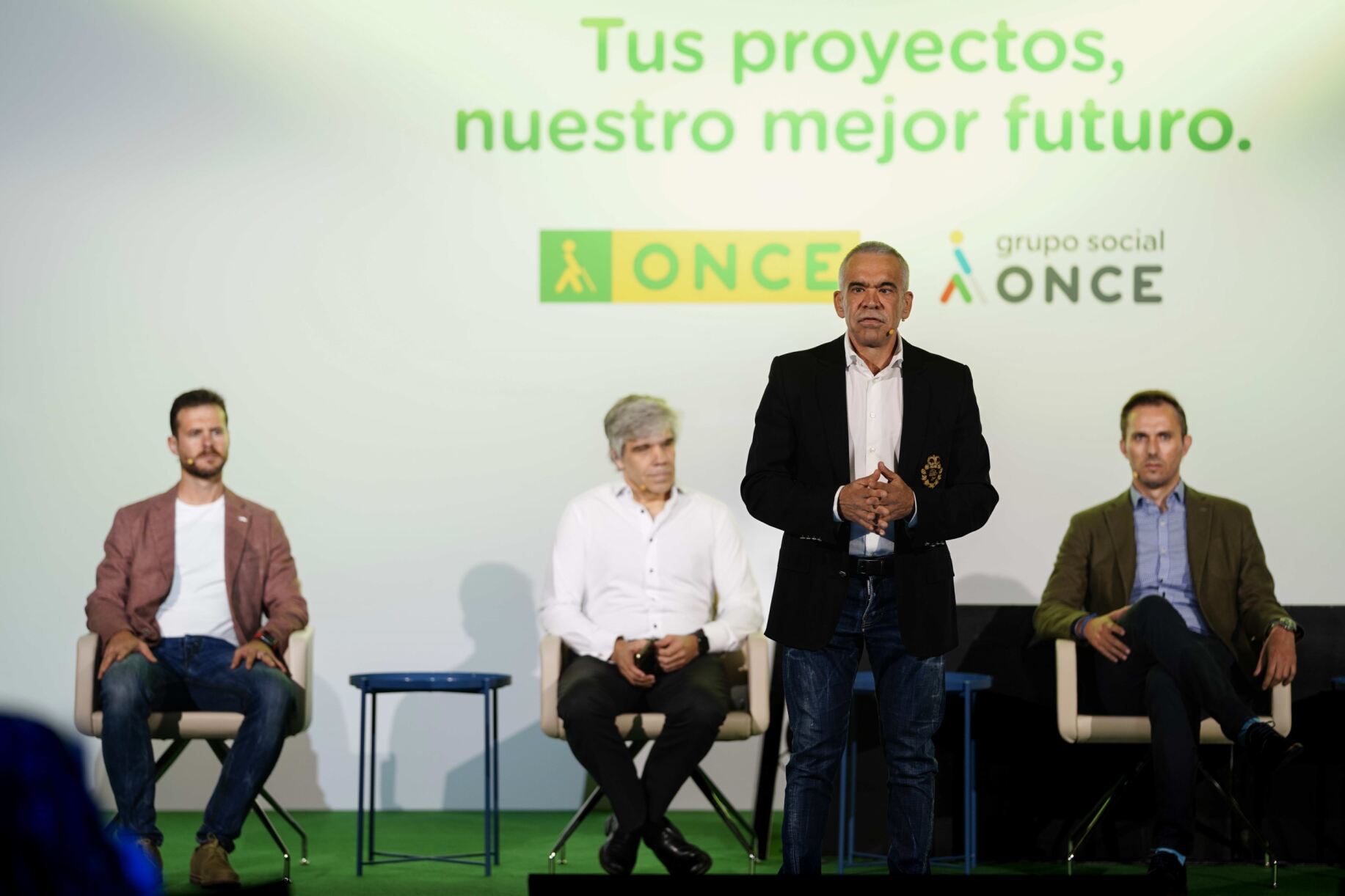 Imagen de un escenario en el que hay un ponente principal, de pie, hablando y tres ponentes al fondo sentados cada uno en una fila. En la parte trasera de la imagen se ve una carátula en grande donde se puede leer