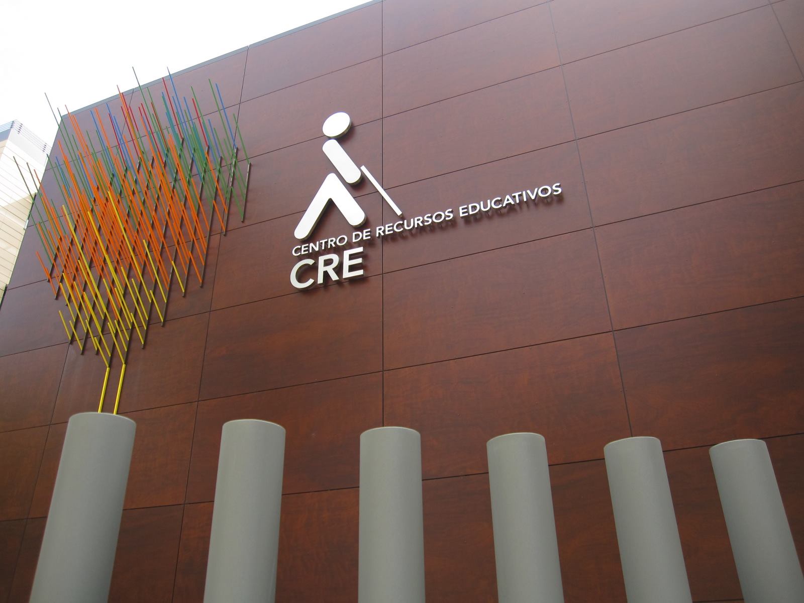 Detalle de la fachada del edificio del CRE en Madrid, donde hay las letras en metal del logo de ONCE, el texto CRE Centro de Recursos Educativos y una instalación de hilos metálicos de colores.