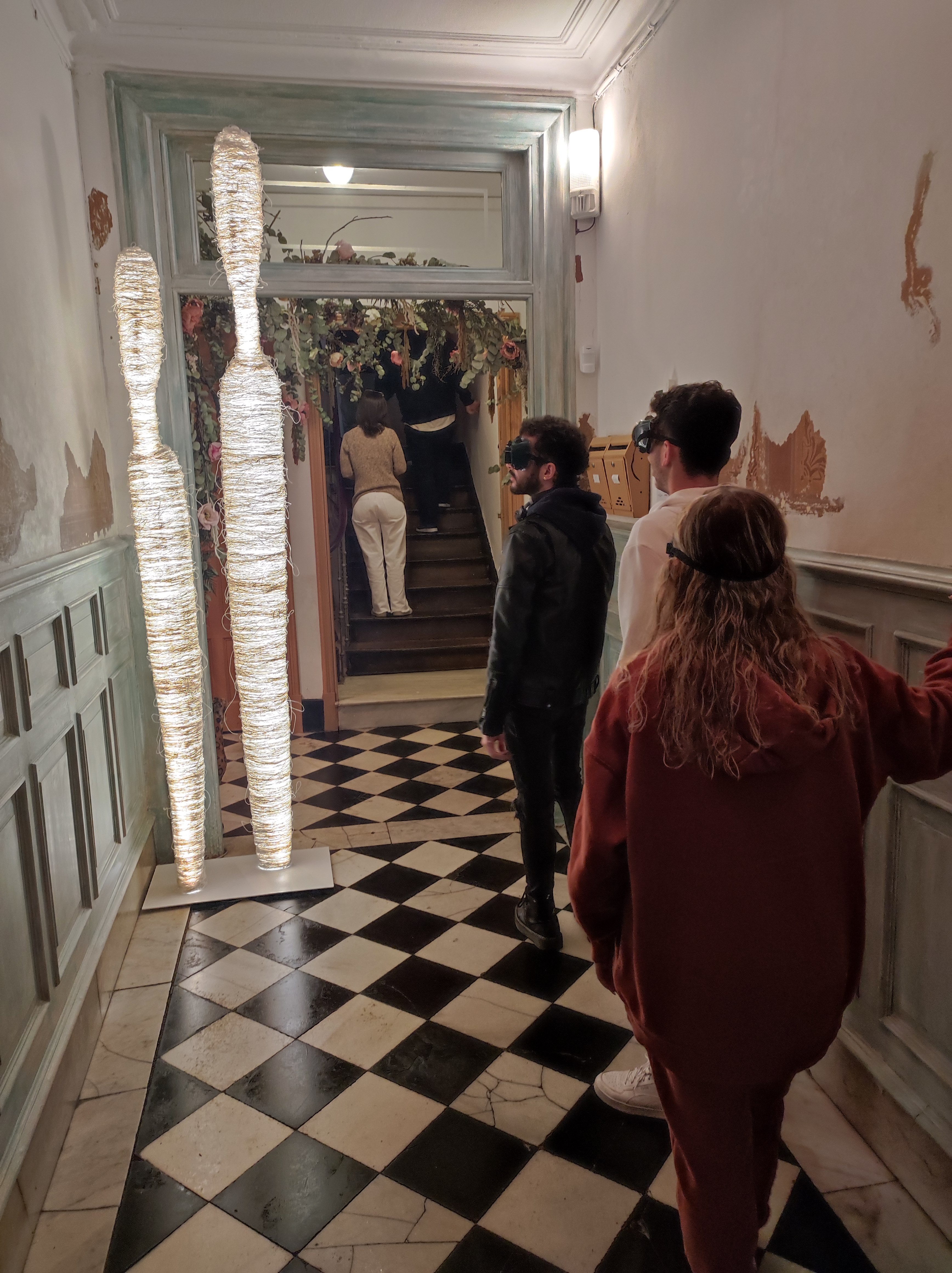 Un grupo de participantes avanzando por un pasillo con gafas de simulación visual, apoyándose en las paredes y guiándose hacia las escaleras decoradas con vegetación.