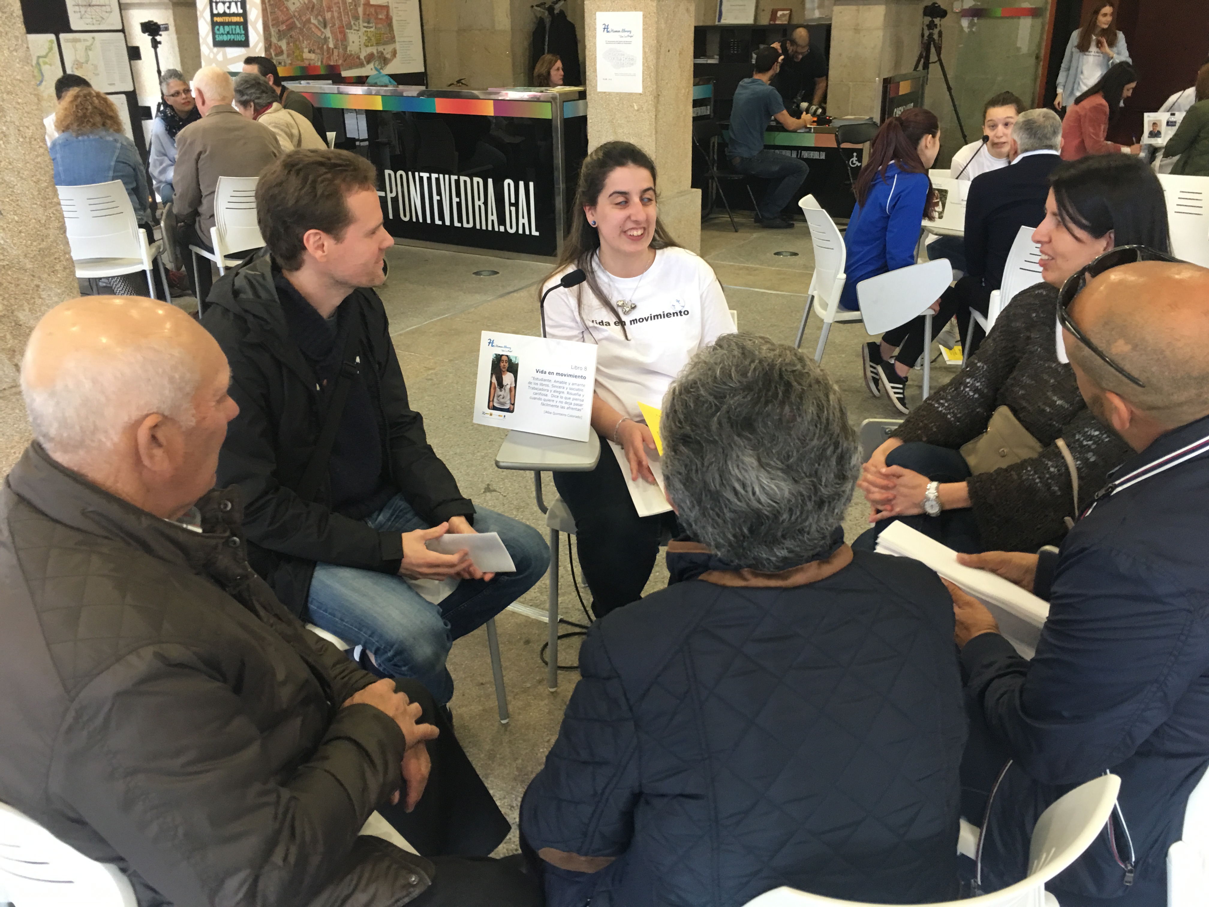 La imagen muestra a un grupo de personas sentadas en sillas, participando en una discusión o reunión. Están en un espacio interior amplio y bien iluminado. Hay cinco personas visibles en la imagen. Una persona está sosteniendo un documento que parece tener texto e imágenes.  Dentro del grupo hay una cartela que pone  “Vida en Movimiento” y la joven que está al lado de la cartela lleva una camiseta blanca con ese mismo mensaje. Todos están enfocados en el documento o en la conversación que están teniendo. El ambiente es informal; están sentados en sillas blancas de plástico. En el fondo, hay otras personas y mesas, lo que indica que es un evento público. Además, hay un mostrador grande con texto visible “PONTEVEDRA.GAL”en vinilo.