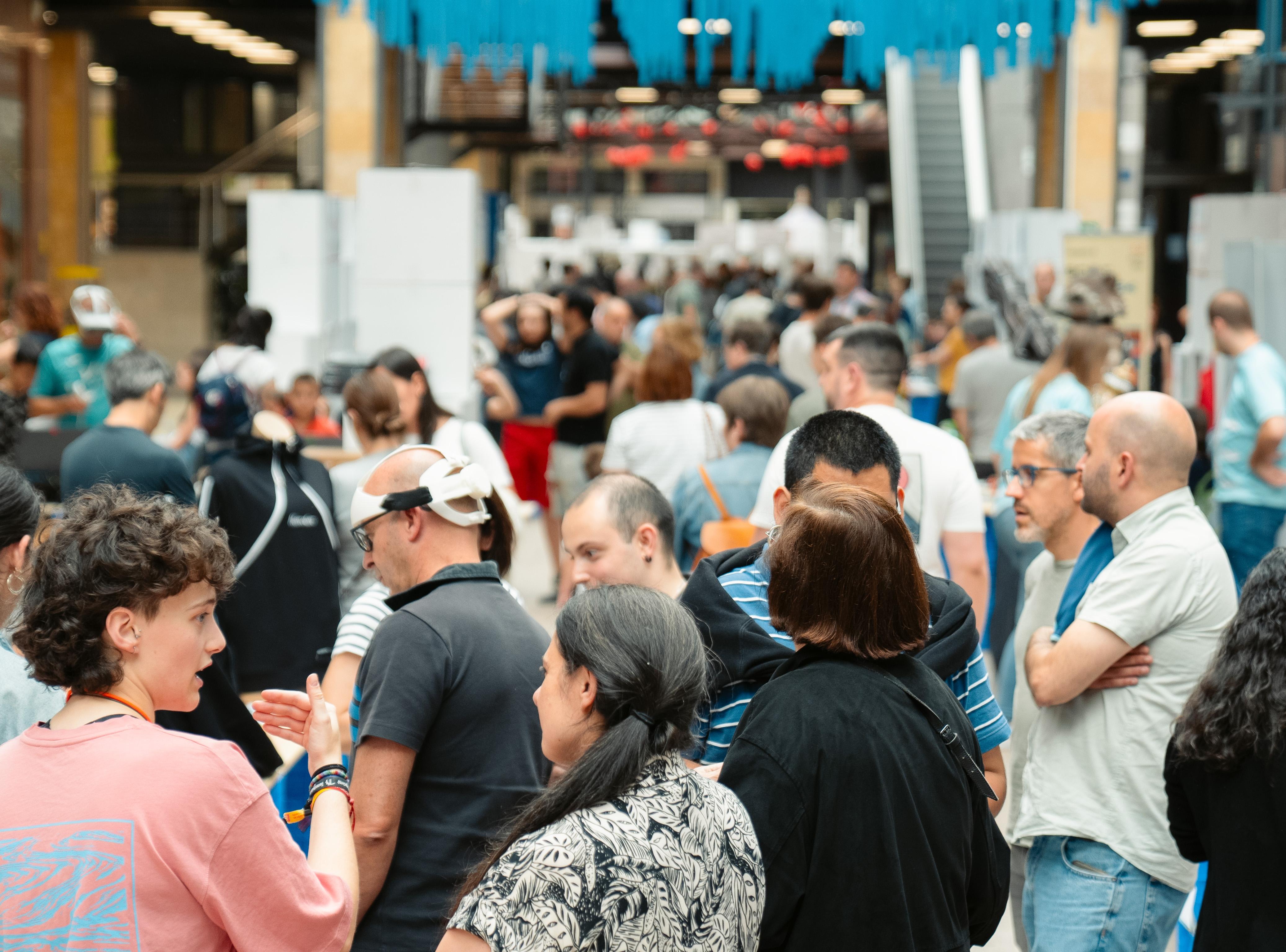 La imagen muestra un evento concurrido en un espacio interior grande. Se observan muchas personas reunidas en grupos pequeños, algunas de las cuales están conversando entre sí. El entorno es la zona de expositores de la Maker Faire Galicia. La multitud incluye una mezcla diversa de personas, tanto hombres como mujeres de diferentes edades. Al fondo, hay una escalera mecánica y decoraciones colgantes en la parte superior, cintas azules. La gente en primer plano está interesada en lo que sucede fuera del marco de la imagen.La atmósfera general es animada y bulliciosa, típica de un evento donde se reúnen muchas personas para intercambiar información, productos o ideas.