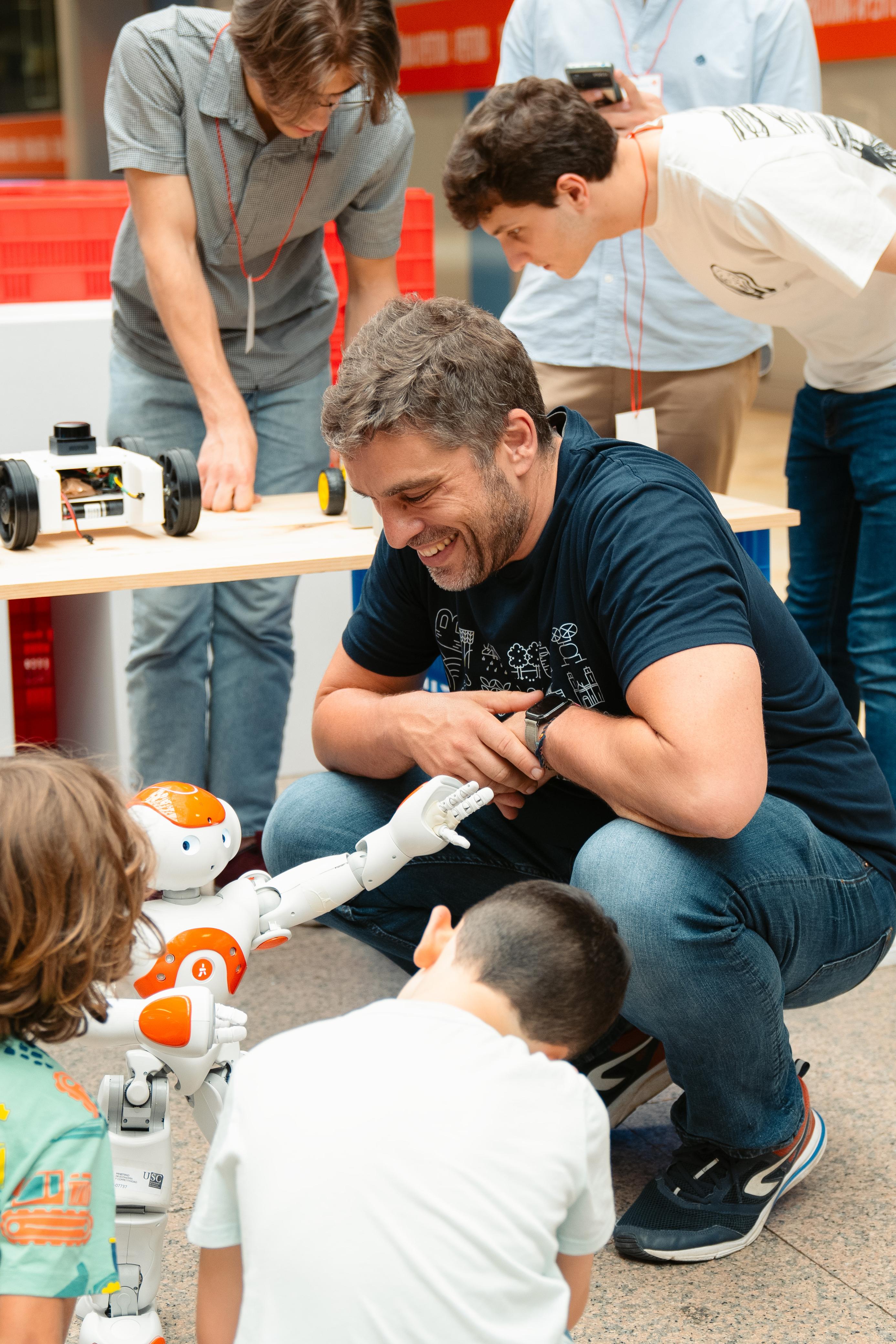 En primer plano, hay un hombre arrodillado sonriendo y participando en la actividad con tres niños. Los niños están concentrados en un pequeño robot humanoide blanco y naranja que está interactuando con ellos. El hombre parece estar guiando o ayudando a los niños mientras observan el robot. Detrás de ellos, hay dos jóvenes adultos, uno de ellos está inclinado sobre una mesa con vehículos robotizados, y el otro está observando atentamente. El entorno es dentro del espacio de exhibición de los expositores de la Maker Faire Galicia, con mesas y material de trabajo.