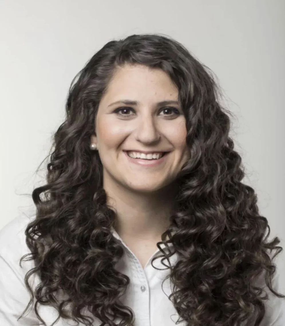 Foto de Natalia Rodriguez. En esta foto Natalia está sonriente y tiene el cabello rizado y voluminoso y lleva una camisa blanca 