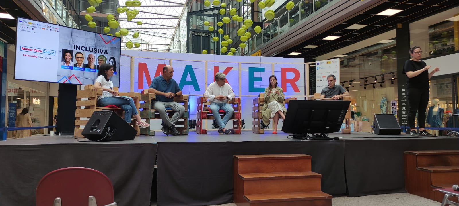 La imagen muestra un escenario en un centro comercial donde se está llevando a cabo un evento titulado "Maker Faire Galicia". En el escenario, hay seis personas sentadas en sillas hechas de palets de madera. Una pantalla grande a la izquierda del escenario muestra información sobre el evento, incluyendo las palabras "INNOVA INCLUSIVA" y la fecha "29 DE XUÑO 16:00 a 18:45". También hay imágenes de algunos de los participantes del evento en la pantalla. Los seis panelistas en el escenario parecen estar involucrados en una discusión. Al lado derecho del escenario, una mujer está haciendo lengua de signos para el público. Encima del escenario hay una decoración con elementos colgantes amarillos, y el fondo muestra el logotipo "MAKER" con letras grandes y coloridas. La audiencia está frente al escenario, aunque no son visibles en la imagen.