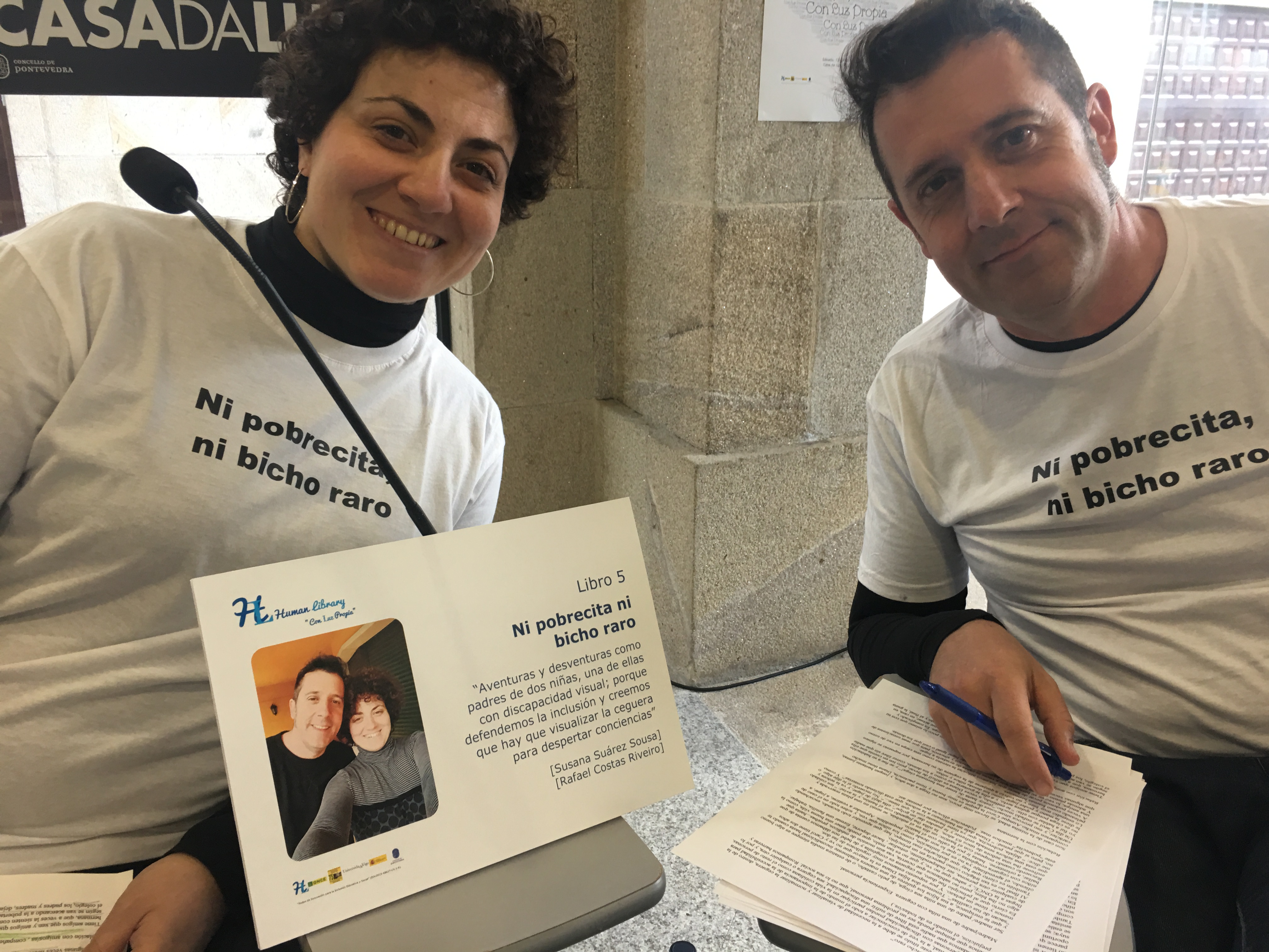 En la imagen salen dos personas, el padre y la madre del Libro humano 5 "Ni pobrecita ni bicho raro". Los dos están sentados sonriendo, llevando puesta una camiseta blanca con el título del libro y entre ellos hay una cartela con el nombre del libro y la descripción "Aventuras y desventuras como padres de dos niñas, una de ellas con discapacidad visual, porque defendemos la inclusión y creemos que hay que visualizar la ceguera para despertar conciencias". Detrás de la cartela y delante de la madre hay un micro y los dos llevan papeles con el guión de la historia.