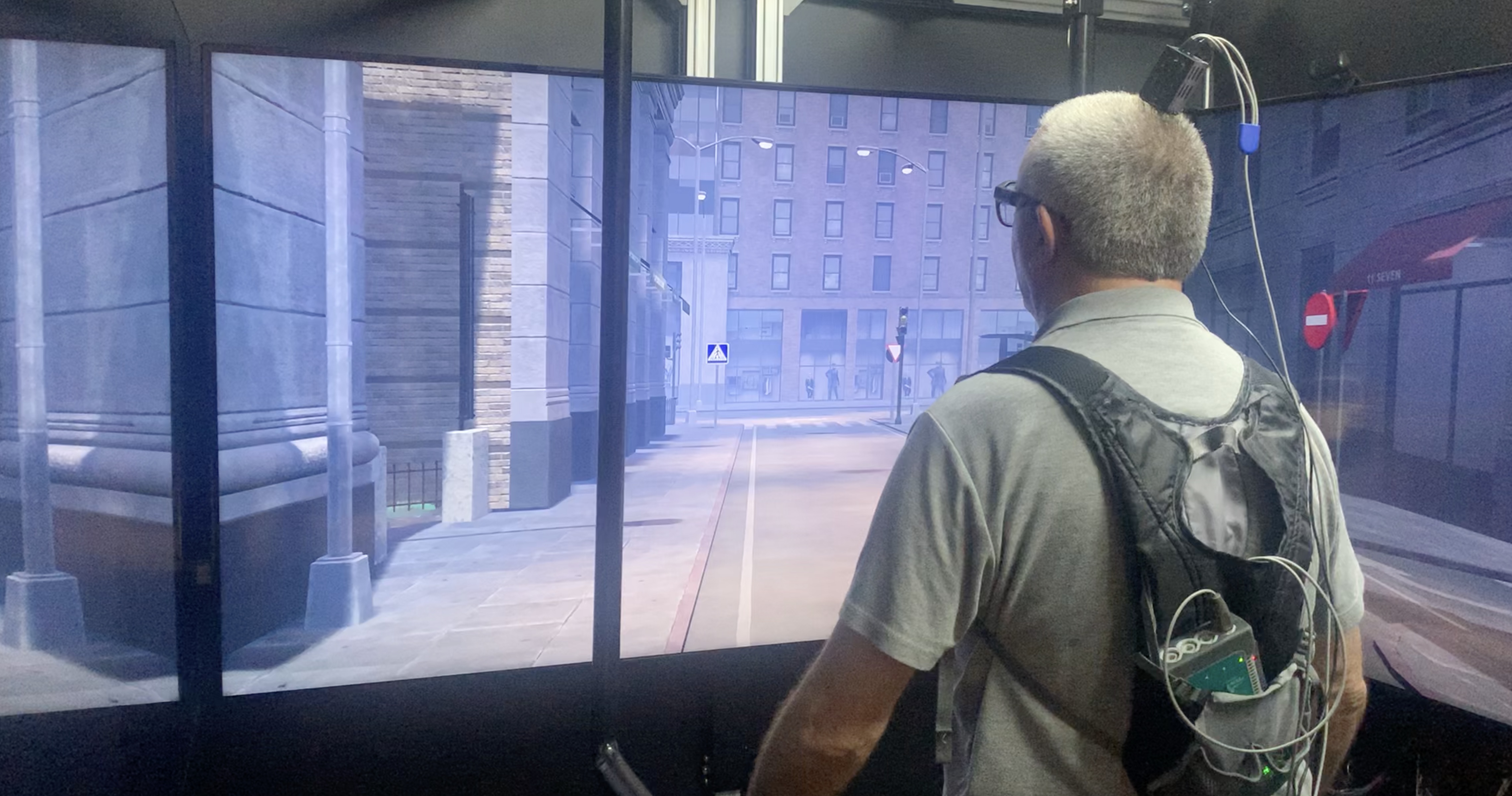 Hombre con casco de realidad virtual mirando hacia una pantalla que cubre todo el espacio. Está inmerso en un simulador de videojuego de realidad virtual que muestra una calle de Nueva York con una intervención urbana. La imagen está capturada desde la parte posterior