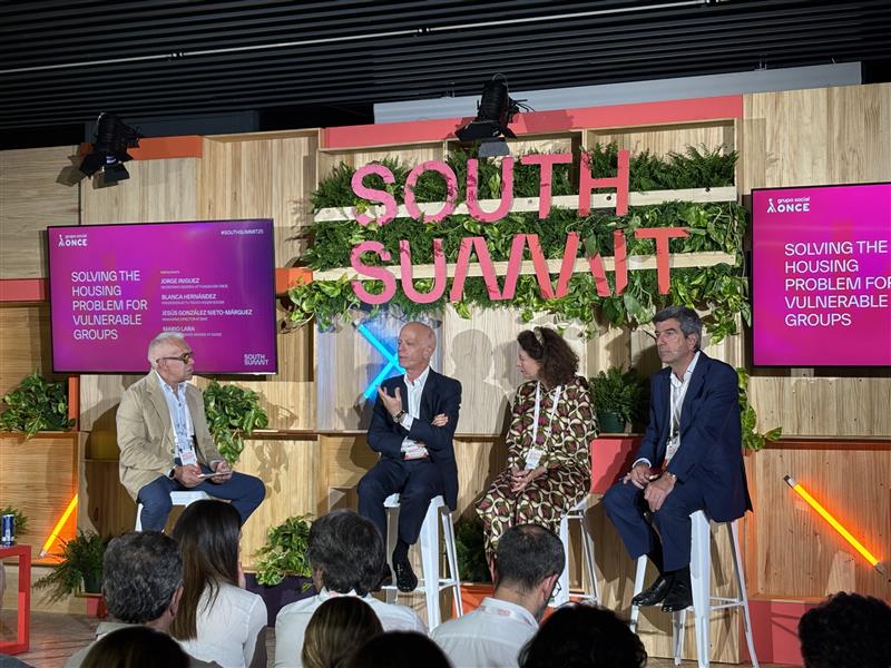 Panel “Solving the housing problem for vulnerable groups”, moderado por Jorge Íniguez en el escenario principal del South Summit. Junto a él, conversan las personas ponentes Blanca Hernández, Jesús González Nieto y Mario Lara sobre innovación social aplicada a soluciones habitacionales dirigidas a colectivos vulnerables. El entorno, decorado con vegetación natural integrada y pantallas digitales con el título del panel, refuerza el enfoque sostenible y humano del encuentro. La escena muestra una audiencia atenta y un debate comprometido en torno a la inclusión desde el ámbito urbano y residencial.