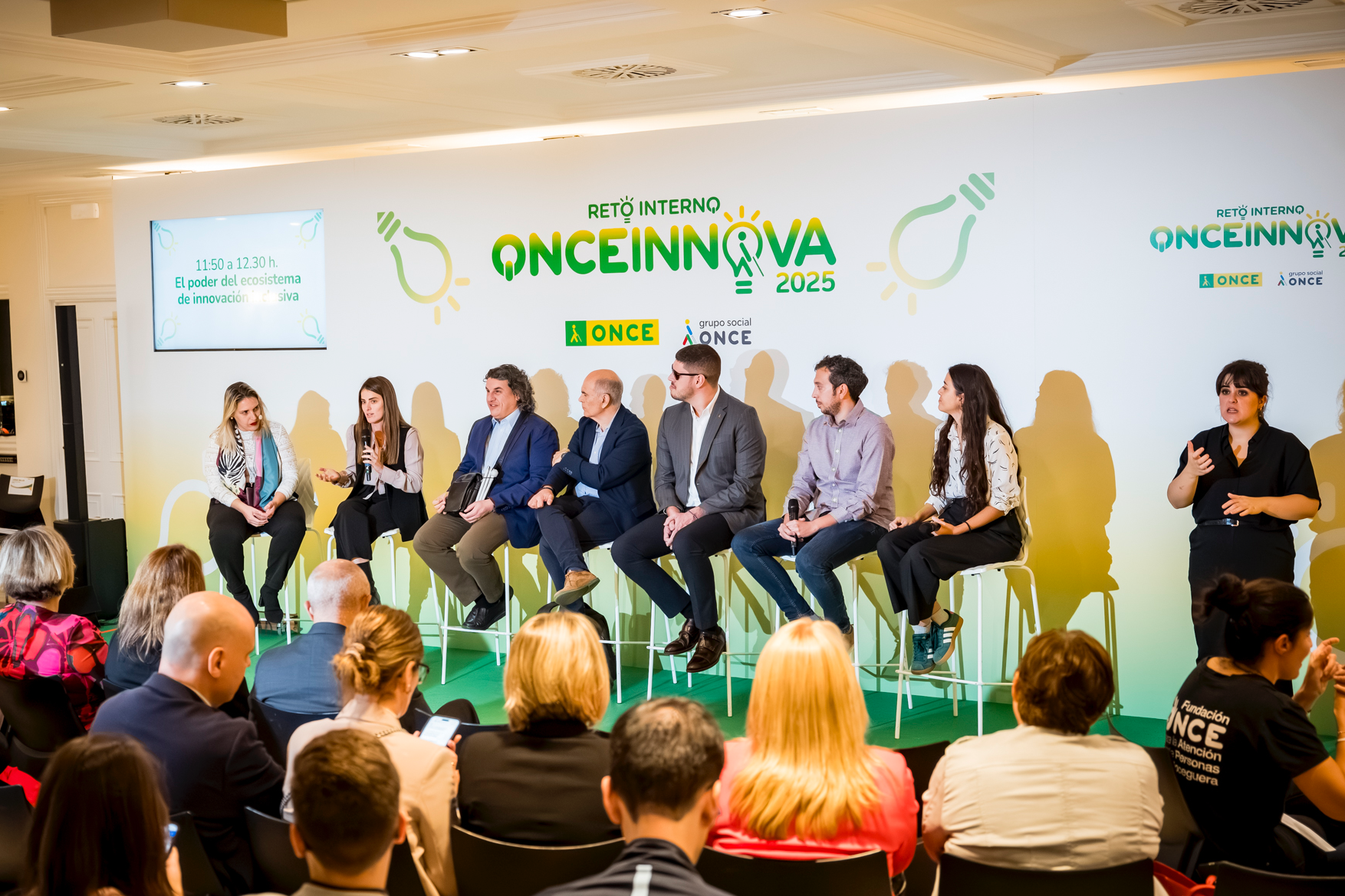 Panel de la final del Reto Interno ONCE Innova 2025. En el escenario aparece un panel de varias personas sentadas en taburetes altos, participando en una conversación o mesa redonda. Todas miran hacia el público o interactúan entre ellas, algunas con micrófono en la mano, en un ambiente formal pero cercano. El fondo del escenario está completamente personalizado con la identidad visual del Reto Interno ONCE Innova 2025, con un diseño en tonos verdes y amarillos, iconos de bombillas y los logotipos de ONCE y Grupo Social ONCE. A la izquierda se ve una pantalla con el título de una de las sesiones del programa, relacionada con el ecosistema de innovación. A la derecha del escenario aparece una intérprete de lengua de signos,