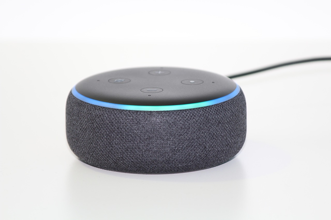 Primer plano de un dispositivo inteligente de tipo altavoz (Amazon Echo Dot con tecnología Alexa), de forma cilíndrica, con acabado textil oscuro. En la parte superior se aprecian los botones de control y el característico aro luminoso azul que indica que el dispositivo está activo.