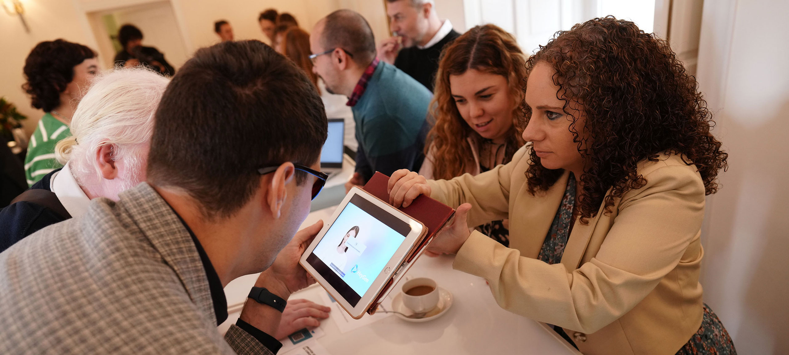 Grupo de personas alrededor de una mesa. Una persona sostiene una tablet que muestra lo que parece ser la foto de perfil de alguien. Sobre la mesa hay varios objetos, incluyendo al menos una taza de café y algunos papeles o cuadernos. La escena parece representar una reunión o encuentro donde se está utilizando tecnología, posiblemente para discusión o presentación.