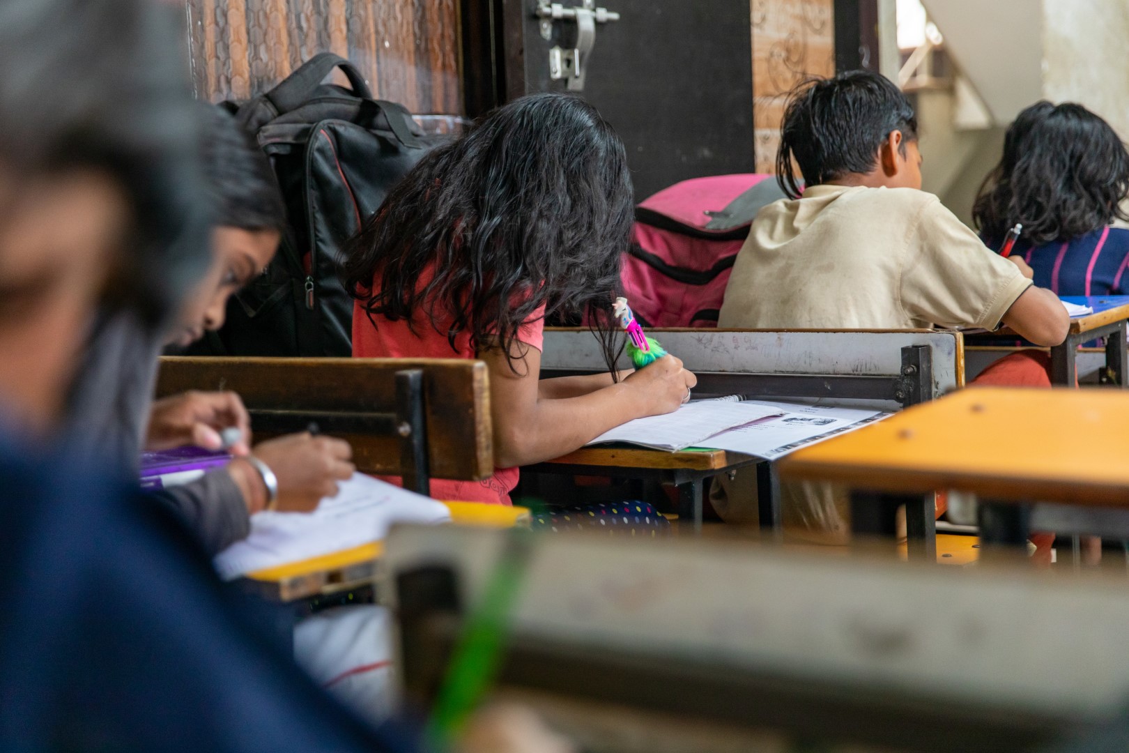 Niños y niñas en una escuela infantil de la India. La foto está tomada desde atrás y se ve a los niños y niñas en sus pupitres realizando tareas. Esta foto pertenece al Proyecto educativo contra el abandono escolar para menores de familias vulnerables de la ONG Itwillbe