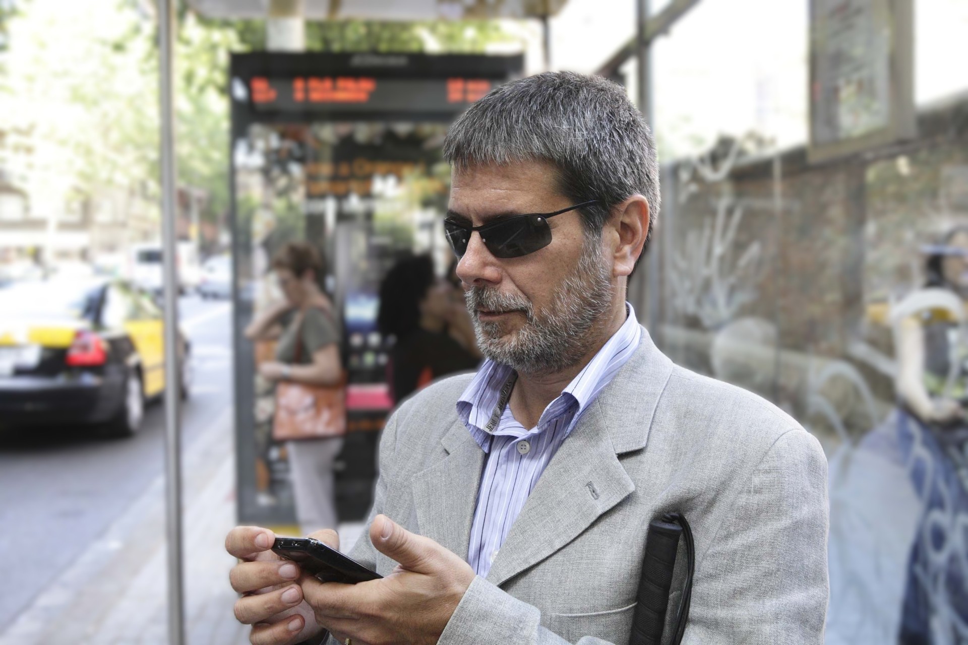 La imagen muestra a Jordi Roig vestido con un traje de color claro y una camisa de rayas. Lleva gafas de sol oscuras y sostiene un teléfono móvil en sus manos, con una expresión de concentración,  lleva también un bastón plegable negro, lo que sugiere que es una persona con discapacidad visual. El fondo está desenfocado y muestra una parada de autobús, de Barcelona, con un panel electrónico con información de horarios. Hay varias personas esperando, algunas de ellas con bolsas o mochilas. También se observan taxis y vehículos en la calle. 
