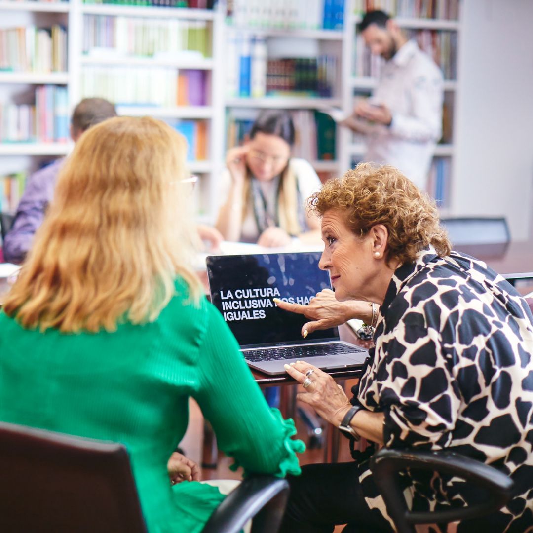 Dos mujeres en una biblioteca con un ordenador que tiene el ZoomText y el texto en el ordenador: la cultura inclusiva nos hace iguales