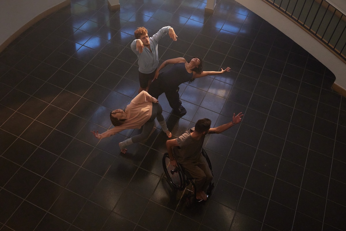 Cuatro personas bailando en un hall rodeados por una escalera de caracol. Uno de ellos va en silla de ruedas