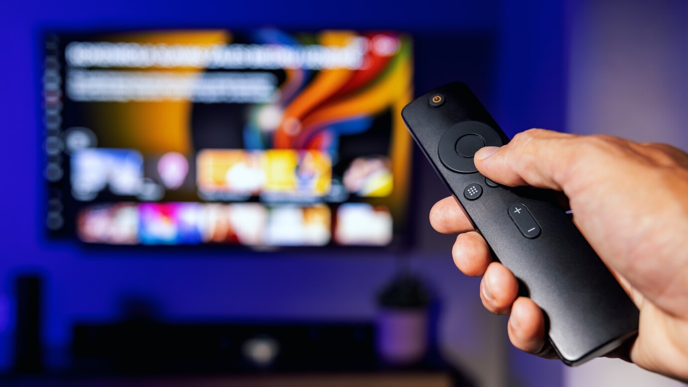 Una mano sujetando un mando de TV, al fondo una pantalla borrosa con aplicaciones de vídeo