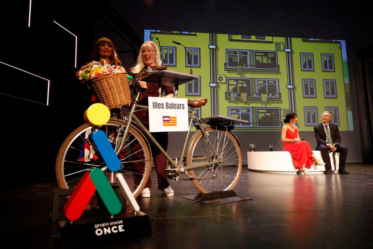 Presentadoras de los Premios Solidarios junto a un atril con forma de bicicleta rotulado con "Illes Balears". En primer plano, figura del Grupo Social ONCE; al fondo, escenario con personas sentadas y una ilustración de edificios