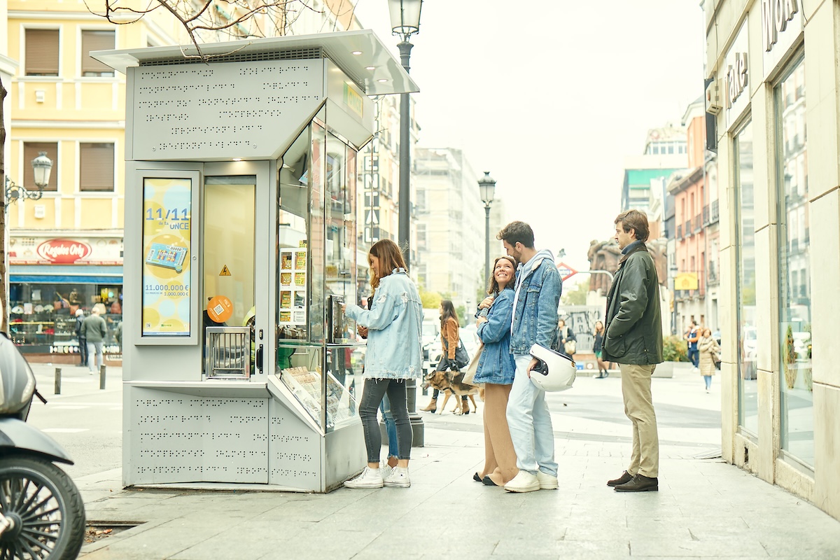 Una fila de personas ante un kiosco de la ONCE en una calle luminosa