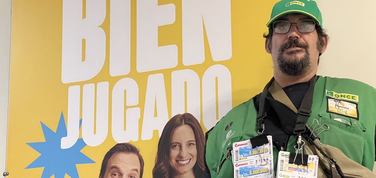 Un vendedor de la ONCE con varios juegos colgados en su chaleco y al fondo un póster que dice 