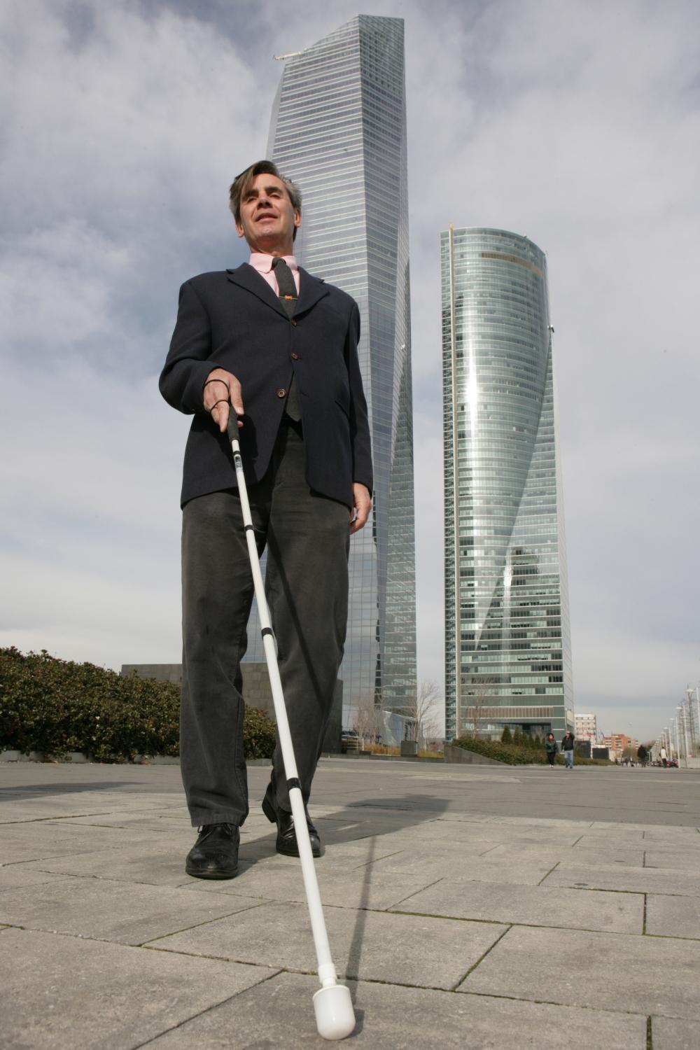 Foto de una persona ciega caminando por la calle, bastón en primer plano, al fondo rascacielos